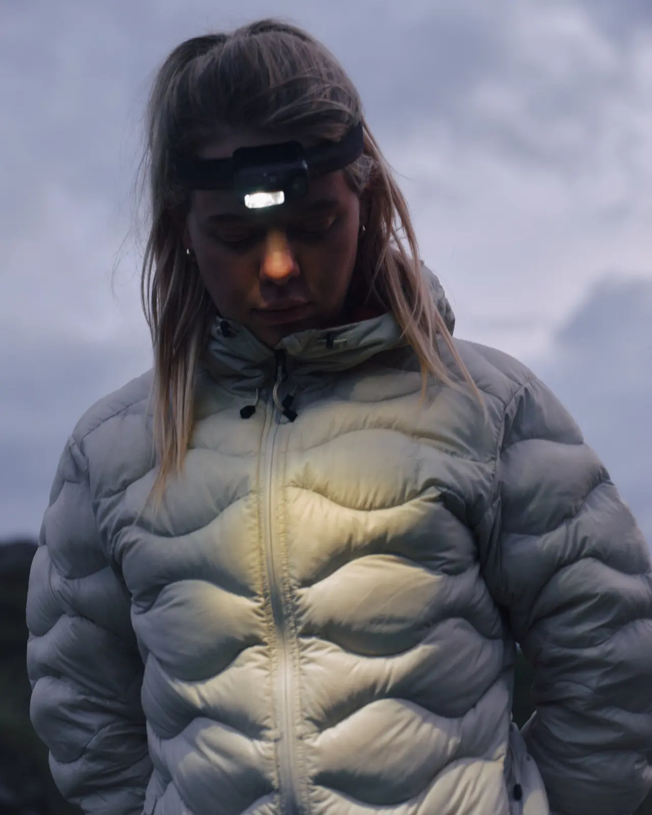 Hiker in a gray puffer jacket looking down while wearing a lit headlamp