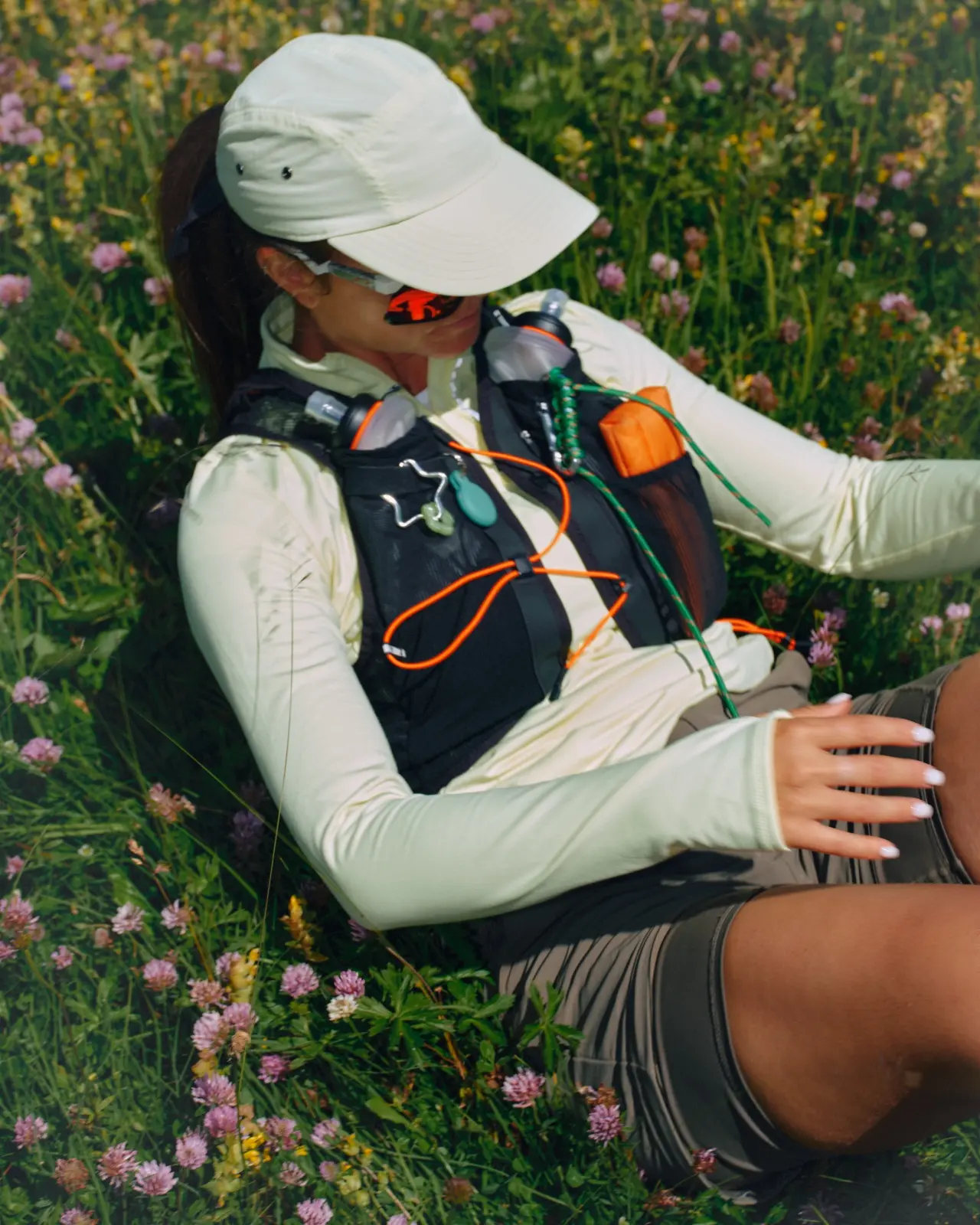 Hiker wearing a white cap and hydration vest resting among alpine wildflowers